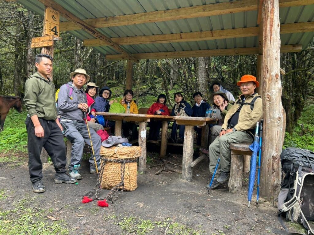 Group of trekkers and guides in safety helmets resting at a wooden trail shelter in a Bhutanese forest
